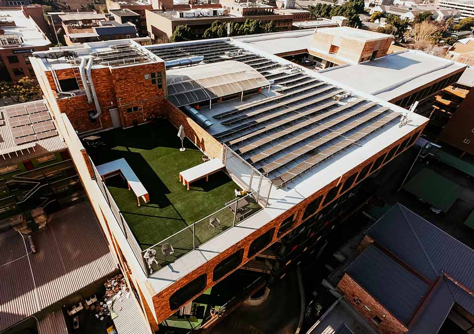 NXT Gen Phase 1: aerial view of the completed brick commercial building, showing the rooftop solar array and the artificial turf roof terrace.